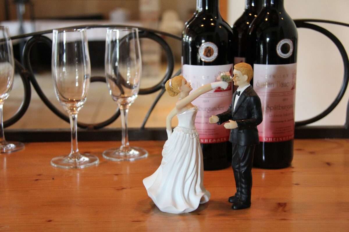 Two wedding cake toppers of a young white man and woman stand in front of wine and glasses